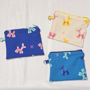 May include: Three small, square fabric pouches with zipper closures. One is navy blue with pink and blue balloon dog prints, another is light blue with blue balloon dog prints, and the third is cream with yellow and pink balloon dog prints.
