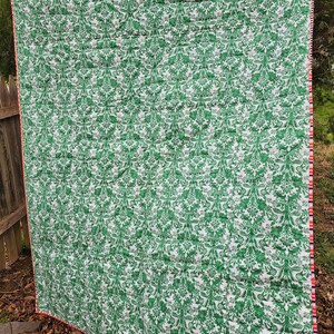 May include: A green and white patterned quilt with a red and white trim. The quilt is folded and lying on the ground.