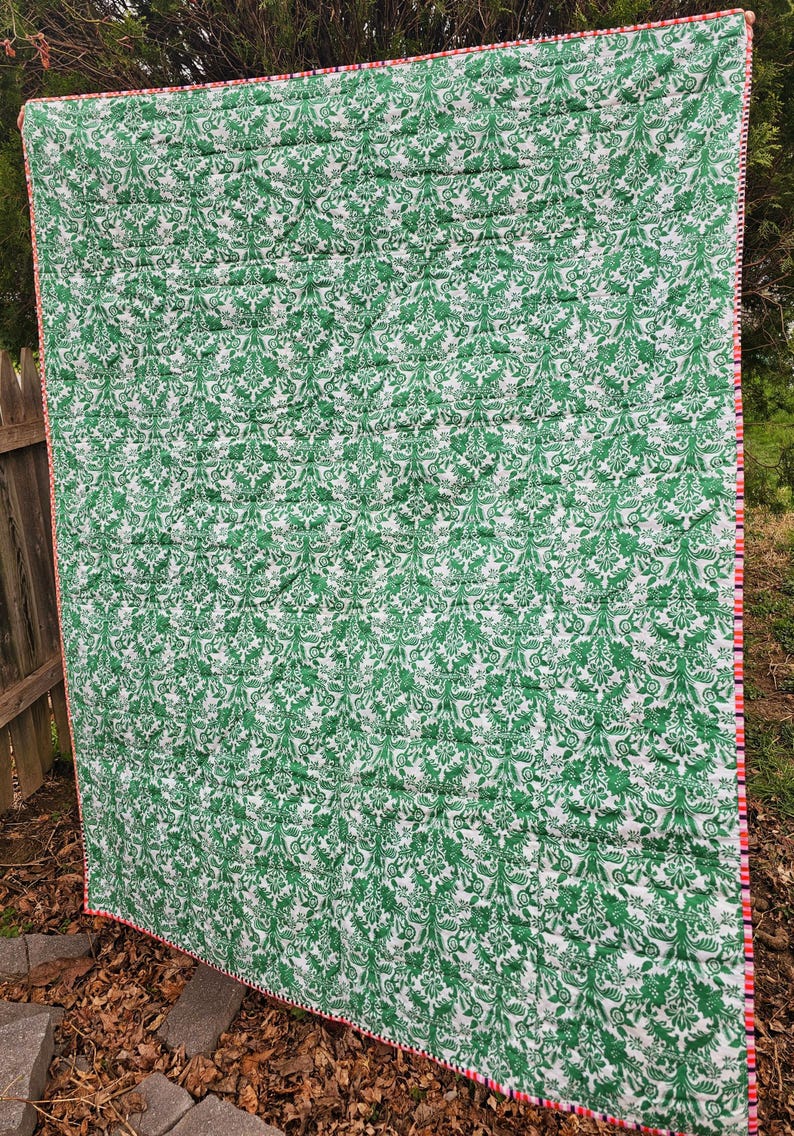 May include: A green and white patterned quilt with a red and white trim. The quilt is folded and lying on the ground.