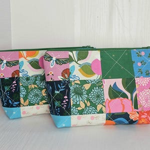 May include: Two patchwork zipper pouches with floral and butterfly patterns. The pouches feature a variety of fabrics in pink, green, blue, and orange, with green zippers. The design is a mix of geometric shapes and botanical motifs.
