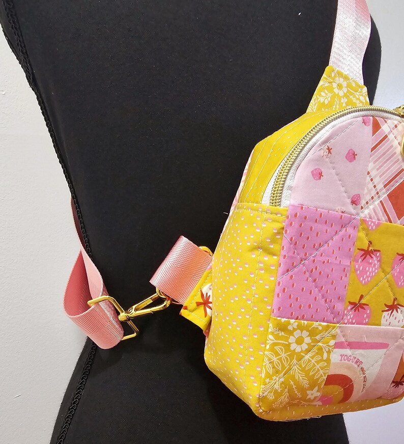 K&ouml;nnte beinhalten: Eine kleine gelbe, rosafarbene und wei&szlig;e gesteppte Umh&auml;ngetasche mit einem goldenen Rei&szlig;verschluss und einem rosafarbenen verstellbaren Trageriemen. Die Tasche hat ein Patchwork-Design mit verschiedenen Mustern, darunter Erdbeeren, Blumen und Punkte.