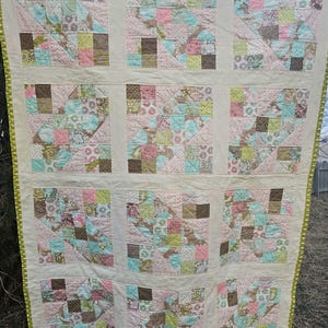 May include: A handmade quilt with a patchwork design. The quilt features squares of various patterns and colours, including pink, brown, green, and blue fabrics. It has a cream-coloured background and a green zigzag border. The quilt is displayed outdoors.