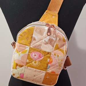 K&ouml;nnte beinhalten: Eine kleine, gesteppte Umh&auml;ngetasche mit Patchwork-Design in Gelb-, Rosa- und Wei&szlig;t&ouml;nen. Die Tasche hat einen Rei&szlig;verschluss und einen verstellbaren braunen Schultergurt.