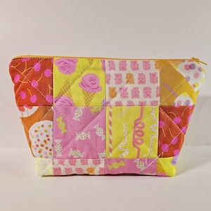 May include: A quilted cosmetic bag with a patchwork design. The bag features squares of fabric in shades of pink, yellow, and orange, with patterns of ice cream cones, candies, and cherries. It has a yellow zipper.