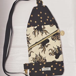May include: Black and cream-colored sling bag with an octopus print and gold star accents. The bag features a black adjustable strap, a gold zipper, and a small zippered pocket. The text "Indie BeBe" is printed on the bottom of the bag.