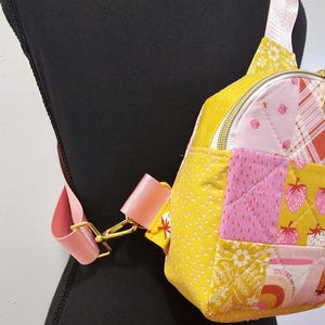 K&ouml;nnte beinhalten: Eine kleine gelbe, rosafarbene und wei&szlig;e gesteppte Umh&auml;ngetasche mit einem goldenen Rei&szlig;verschluss und einem rosafarbenen verstellbaren Trageriemen. Die Tasche hat ein Patchwork-Design mit verschiedenen Mustern, darunter Erdbeeren, Blumen und Punkte.