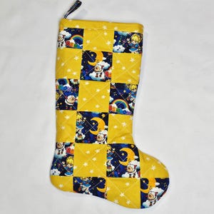 May include: A quilted Christmas stocking with a yellow background and patchwork squares. The squares feature a cartoon character with a crescent moon, rainbow, and stars. The stocking has a hanging loop.