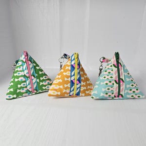 May include: Three triangular zippered pouches with colorful geometric patterns. The pouches are made of fabric and have a pink zipper. The pouches are lined up in a row from left to right: green, yellow, and blue.