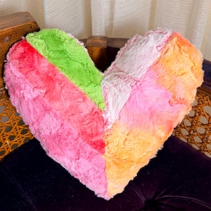 May include: A heart-shaped pillow made of soft, plush faux fur fabric. The pillow features a vibrant, multi-colored chevron pattern with distinct sections of bright lime green, hot pink, light pink, and warm orange hues throughout the surface.