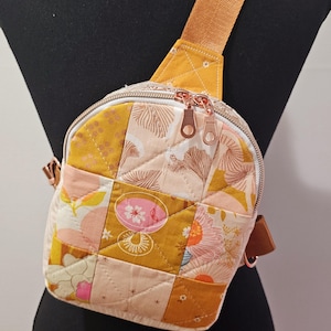 May include: A small, quilted crossbody bag with a patchwork design in shades of yellow, pink, and white. The bag has a zipper closure and a brown adjustable strap.
