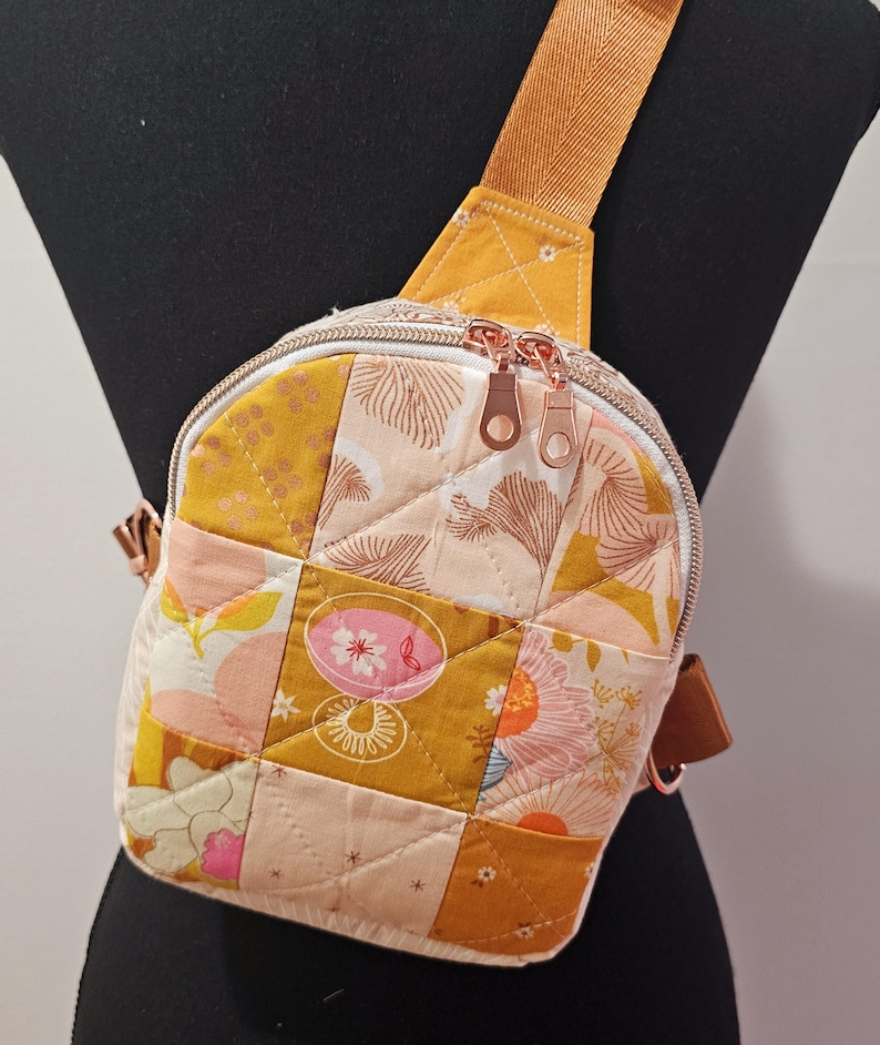 K&ouml;nnte beinhalten: Eine kleine, gesteppte Umh&auml;ngetasche mit Patchwork-Design in Gelb-, Rosa- und Wei&szlig;t&ouml;nen. Die Tasche hat einen Rei&szlig;verschluss und einen verstellbaren braunen Schultergurt.