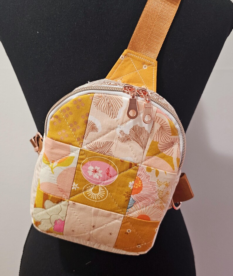 K&ouml;nnte beinhalten: Eine kleine, gesteppte Umh&auml;ngetasche mit Patchwork-Design in Gelb-, Rosa- und Wei&szlig;t&ouml;nen. Die Tasche hat einen Rei&szlig;verschluss und einen verstellbaren braunen Schultergurt.
