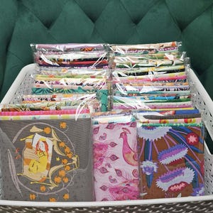 May include: A white basket filled with stacks of fabric. The fabrics are in clear plastic packaging. The fabrics have various patterns, including floral, geometric, and illustrated designs. The background is a green, tufted surface.