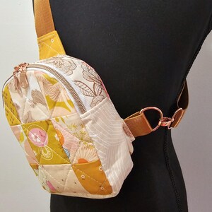 K&ouml;nnte beinhalten: Eine kleine gesteppte Umh&auml;ngetasche mit Blumendruck und einem braunen Riemen. Die Tasche hat einen Rei&szlig;verschluss und eine goldene Metallschnalle.