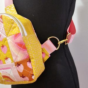 K&ouml;nnte beinhalten: Eine kleine gelbe und rosafarbene gesteppte Umh&auml;ngetasche mit einem goldenen Rei&szlig;verschluss und einem rosafarbenen Riemen. Die Tasche hat ein Patchwork-Design mit Erdbeeren und Regenb&ouml;gen.
