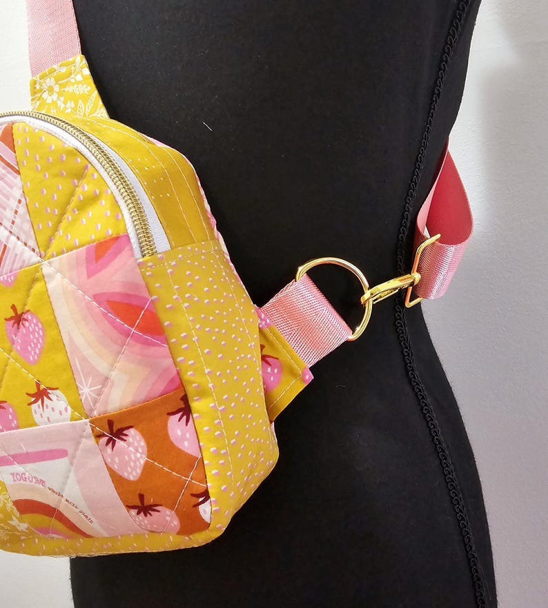 K&ouml;nnte beinhalten: Eine kleine gelbe und rosafarbene gesteppte Umh&auml;ngetasche mit einem goldenen Rei&szlig;verschluss und einem rosafarbenen Riemen. Die Tasche hat ein Patchwork-Design mit Erdbeeren und Regenb&ouml;gen.
