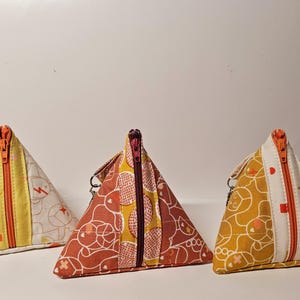 May include: Three triangular zippered pouches made of fabric. The pouches are in different colors and patterns. The first pouch is white with orange and yellow geometric patterns. The second pouch is brown with orange and white geometric patterns. The third pouch is yellow with orange and white geometric patterns.