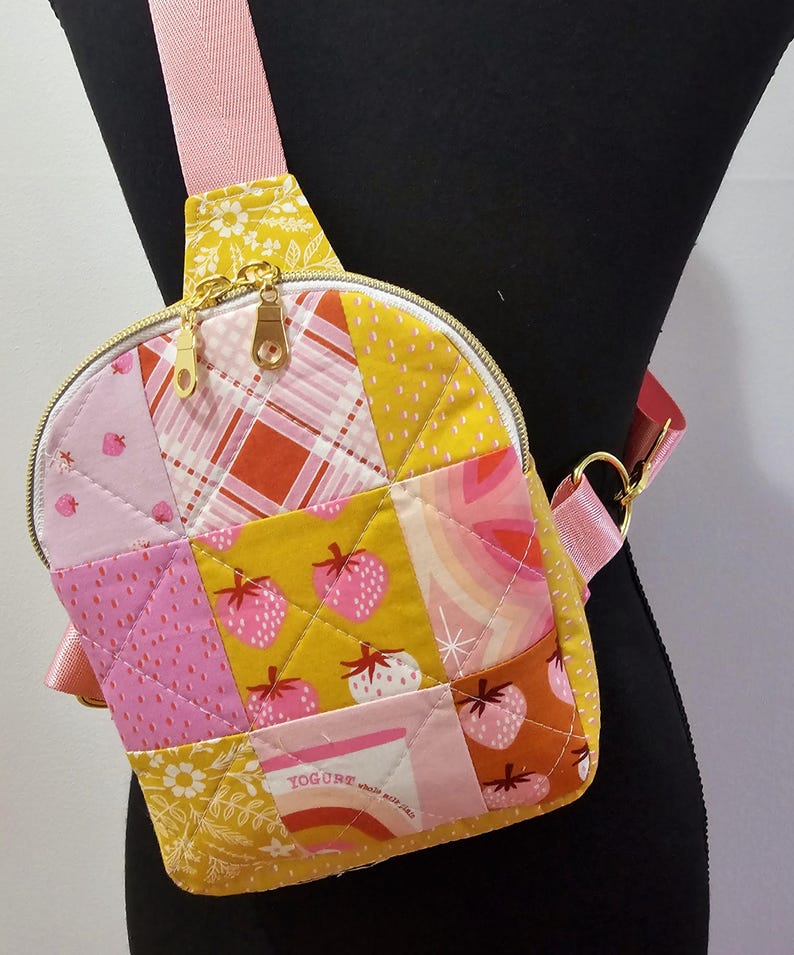 K&ouml;nnte beinhalten: Eine kleine, gesteppte, gelbe und rosafarbene Umh&auml;ngetasche mit Rei&szlig;verschluss. Die Tasche besteht aus Patchwork-Stoff mit verschiedenen Mustern, darunter Erdbeeren, Karos und Blumenmuster. Die Tasche hat einen goldenen Rei&szlig;verschluss und einen rosafarbenen Riemen.