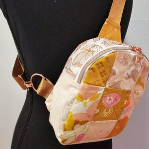 K&ouml;nnte beinhalten: Eine kleine, gesteppte Umh&auml;ngetasche mit Patchwork-Design in Rosa-, Gelb- und Wei&szlig;t&ouml;nen. Die Tasche hat einen Rei&szlig;verschluss und einen ros&eacute;goldenen Riemen.