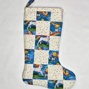 May include: A quilted Christmas stocking with a patchwork design. The pattern features cartoon characters, rainbows, and unicorns on a white background with red and gold stars. A blue loop is attached at the top.