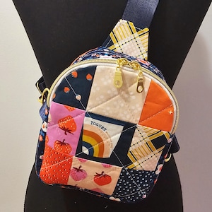 May include: A quilted patchwork crossbody bag with a gold zipper and a navy blue strap. The bag features a variety of colorful patterns, including strawberries, rainbows, and plaid. The bag is small and compact, perfect for carrying essentials.