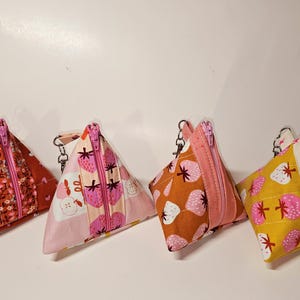 May include: Four small triangular zippered pouches with different strawberry patterned fabric. The pouches are pink, red, orange, and yellow with white accents. Each pouch has a silver zipper and a silver chain.