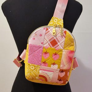 May include: A pink and yellow patchwork fabric crossbody bag with a gold zipper and a strap. The bag has a quilted design and features a strawberry print and the text "Yogurt with granola".