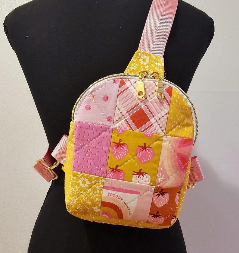 K&ouml;nnte beinhalten: Eine rosa und gelbe Patchwork-Umh&auml;ngetasche mit einem goldenen Rei&szlig;verschluss und einem Trageriemen. Die Tasche hat ein gestepptes Design und zeigt einen Erdbeer-Druck und den Text "Joghurt mit Granola".