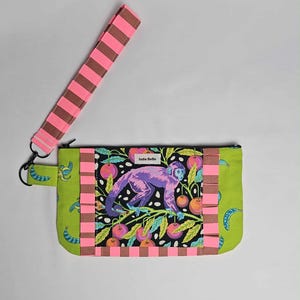 May include: A rectangular clutch with a wrist strap. The clutch features a monkey design on a black background with pink and orange accents. The wrist strap is pink and brown striped. The brand name "Indie Belle" is visible on the clutch.