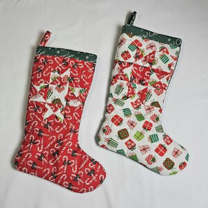 May include: Two Christmas stockings. One is red with white candy canes and a white star design. The other is white with red and green gift boxes. Both have green trim and hanging loops. Festive holiday decor.