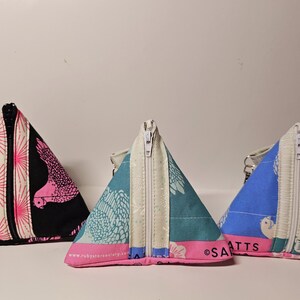 May include: Three triangular zippered pouches with different patterns. The pouch on the left is black with pink and white accents and a pink bird design. The middle pouch is teal with white and pink accents and a white bird design. The pouch on the right is blue with white and pink accents and a white bird design.