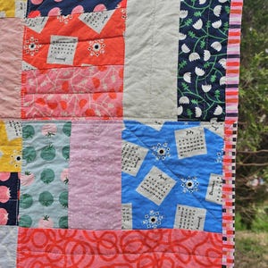 May include: A colorful quilt with a patchwork design featuring various patterns, including floral, geometric, and calendar prints. The quilt is edged with a pink and white striped fabric.