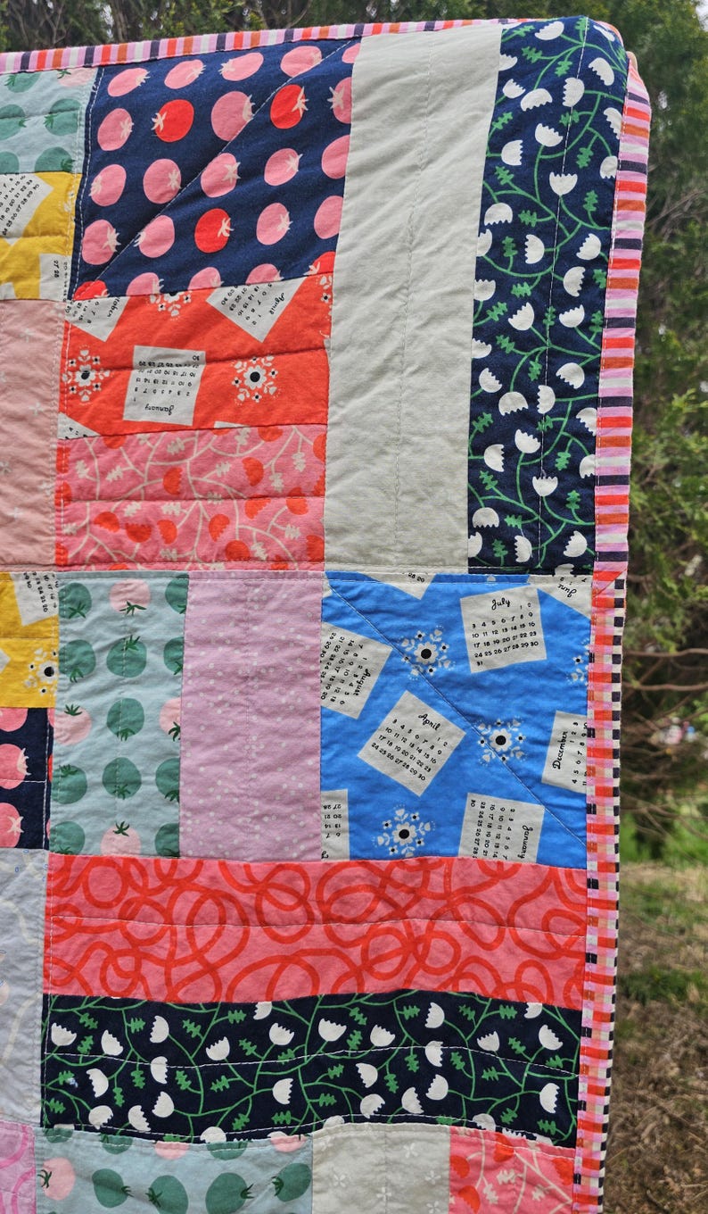 May include: A colorful quilt with a patchwork design featuring various patterns, including floral, geometric, and calendar prints. The quilt is edged with a pink and white striped fabric.