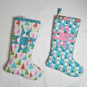 May include: Two Christmas stockings. One is white with colorful trees and a blue star pattern. The other is light blue with snowmen and a pink star pattern. Both have a loop for hanging.