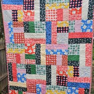 May include: A colorful patchwork quilt with a variety of patterns, including polka dots, floral designs, and geometric shapes. The quilt is made up of squares and rectangles in shades of pink, red, yellow, green, blue, and white.