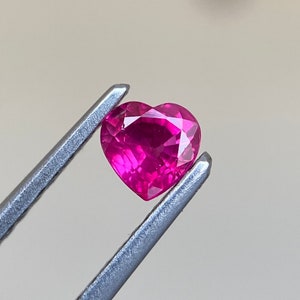 Op de afbeelding: Een hartvormige robijn edelsteen vastgehouden door een pincet. De robijn is diep roze van kleur en heeft een facetgeslepen vorm.