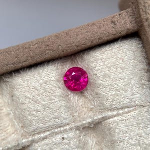 Puede incluir: Una gema rosa vibrante y redonda, probablemente un rubí, colocada sobre una tela suave de color blanco roto. Se destaca la superficie pulida y el color intenso de la gema. El entorno parece ser una exhibición de joyas.