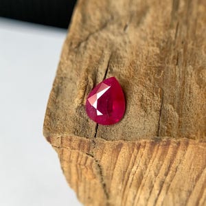 Può includere: Una gemma di rubino sfaccettata a forma di pera. La gemma è di un colore rosso intenso e vibrante con molteplici sfaccettature che riflettono la luce. È posta su una superficie di legno marrone chiaro strutturata, creando un contrasto di colore e consistenza.