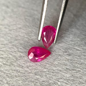 Puede incluir: Dos rubíes facetados en forma de pera. Uno es sujetado por unas pinzas, el otro descansa debajo. Las gemas muestran un color rojo intenso y vibrante sobre un fondo gris texturizado. Estas piedras preciosas son ideales para la fabricación de joyas.