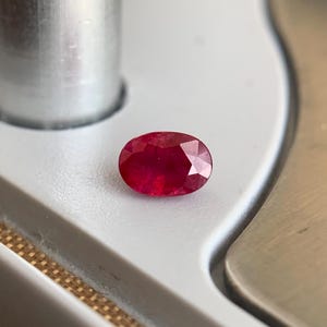 Può includere: Un rubino ovale sfaccettato, di un rosso intenso. La gemma è posizionata su una superficie bianca. Le sfaccettature del rubino riflettono la luce. Lo sfondo è sfocato, evidenziando il colore e la forma della pietra preziosa.