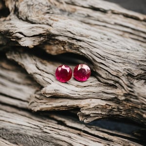 Può includere: Due orecchini rotondi con gemme rosse brillanti e sfaccettate. Sono posizionati su uno sfondo di legno alla deriva grigio e invecchiato, creando un contrasto di trame e colori. Le gemme sono il punto focale, mostrando il loro colore e taglio vivaci.