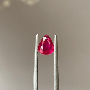 Puede incluir: Un rubí en forma de pera, sostenido por unas pinzas plateadas. La gema de color rojo intenso está facetada para realzar su brillo. El fondo es de un tono suave y neutro, que resalta el color y la forma del rubí.
