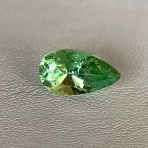 Puede incluir: Una gema facetada en forma de pera con un vibrante tono verde. La gema muestra tonos verdes claros y amarillos, con una superficie pulida que refleja la luz. La gema se encuentra sobre un fondo neutro.