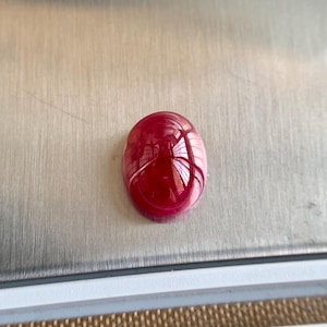 May include: A polished, oval-shaped, deep red gemstone. The gem has a high luster, reflecting light and creating a vibrant appearance. The stone is set against a silver surface, highlighting its color and shape.