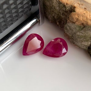 Könnte beinhalten: Zwei birnenförmige Rubingesteine, jeder mit einer tief roten Farbe und einem facettierten Schliff. Die Edelsteine ​​werden auf einer weißen Oberfläche präsentiert.