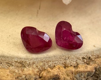 May include: Two heart-shaped ruby gemstones, each with a deep red colour and a faceted cut. The gemstones are displayed on a light brown wooden surface.