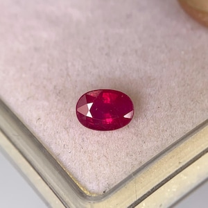 Könnte beinhalten: Ein facettierter, oval geformter Rubin-Edelstein. Der Rubin hat eine tiefe, leuchtende rote Farbe und ist vor einem hellrosa Hintergrund platziert. Der Edelstein wird in einer transparenten Kunststoffbox präsentiert.