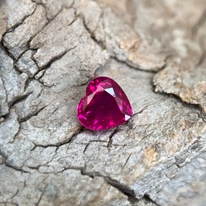 Pode incluir: Uma gema de rubi em forma de coração com uma tonalidade rosa profunda. A gema é colocada contra um fundo áspero e texturizado.