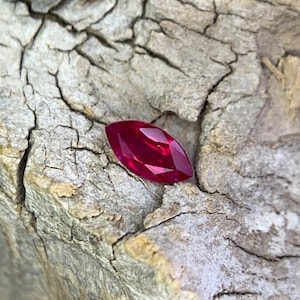 May include: A deep red marquise-shaped gemstone on a rough, textured, grey background.