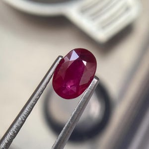 Può includere: Un rubino sfaccettato di forma ovale, tenuto da pinzette d'argento. Il rubino è di un colore rosso intenso e saturo, con riflessi interni visibili. Le pinzette sono a fuoco, evidenziando la chiarezza e la brillantezza della gemma. Lo sfondo è sfocato.